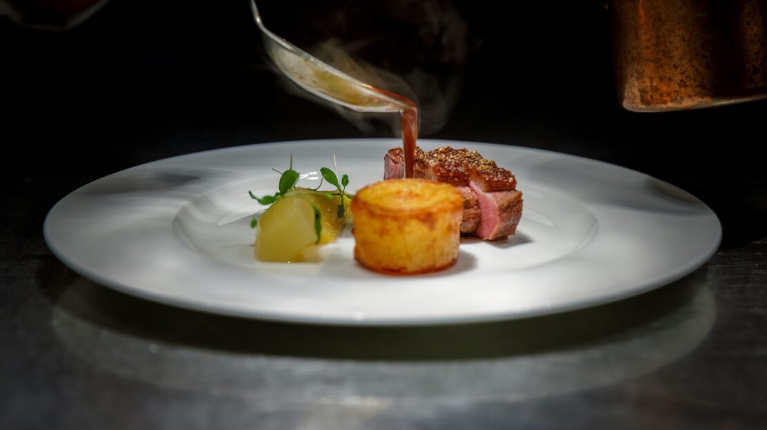 Chef pouring sauce over a plated steak entrée with a gratin and pear garnish at a fine dining restaurant.